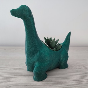 Op de afbeelding: Een groene keramische dinosaurus plantenpot met een kleine succulent plant erin. De dinosaurus staat op een licht houten oppervlak.