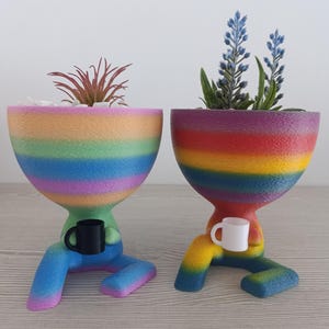 Pode incluir: Dois vasos de plantas com cores de arco-íris em forma de pessoas, cada um segurando uma xícara pequena. Um vaso tem uma xícara preta e o outro tem uma xícara branca. Ambos os vasos têm pequenas plantas crescendo dentro.