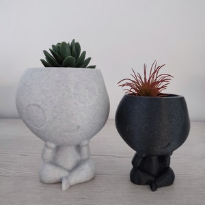 Könnte beinhalten: Zwei kleine, weiße und schwarze Keramik-Pflanztöpfe in Form von Menschen, die in meditativer Pose sitzen. Der weiße Topf hat eine grüne Sukkulente drin und der schwarze Topf eine rote Sukkulente.