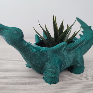 May include: A teal green dinosaur planter with a spiky back and a small succulent plant growing inside.