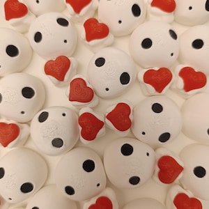 Puede incluir: Una colección de figuritas de fantasmas de cerámica blanca con ojos negros y decoraciones en forma de corazón rojo. Los fantasmas están dispuestos en un patrón aleatorio sobre una superficie blanca.