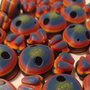 Puede incluir: Colección de objetos coloridos impresos en 3D en varias formas, incluyendo esferas redondeadas y formas orgánicas. Los objetos presentan una vibrante combinación de colores arcoíris con bandas de rojo, naranja, amarillo, verde y azul. Cada pieza tiene un agujero.