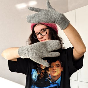 May include: A pair of gray knitted mittens with a matching gray knitted hat. The mittens are made of a soft, textured yarn. The person in the image is wearing a pink beanie, black glasses, and a black t-shirt with a graphic print.