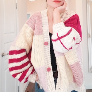 Könnte beinhalten: Eine grob gestrickte Strickjacke mit Patchwork-Design aus cremefarbenen, hellrosa und dunkelrosa Blöcken. Die Ärmel zeigen horizontale rote und weiße Streifen, während die Vorderseite mit zwei runden, strukturierten rosa Knöpfen geschlossen wird.