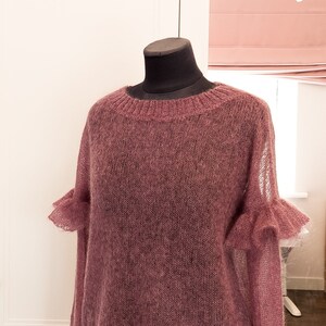 May include: A dusty rose-colored long-sleeve sweater crafted from a sheer, fuzzy knit material. The garment features a ribbed crew neckline and decorative ruffled cuffs on the sleeves, providing a textured appearance throughout the entire piece.