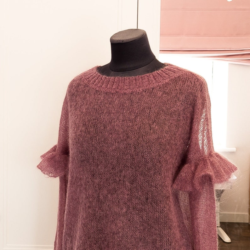 May include: A dusty rose-colored long-sleeve sweater crafted from a sheer, fuzzy knit material. The garment features a ribbed crew neckline and decorative ruffled cuffs on the sleeves, providing a textured appearance throughout the entire piece.