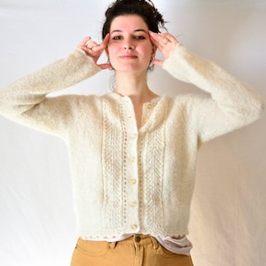 May include: Cream-coloured, button-down cardigan sweater. The sweater has a textured knit pattern with a scalloped hemline. The long-sleeved sweater is made of a soft, fuzzy material.
