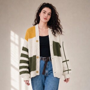May include: A chunky knit cardigan featuring a color-blocked design in cream, mustard yellow, and olive green. The garment has a V-neckline, long sleeves with striped and grid patterns, and three round buttons down the front closure.