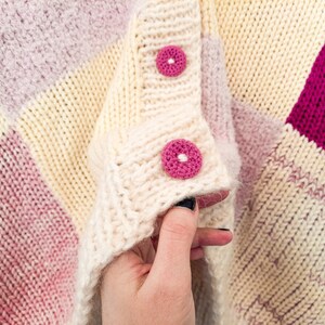 May include: A chunky knit cardigan featuring a color-blocked pattern in cream, soft pink, and vibrant magenta. The garment includes a thick ribbed placket with two round, textured magenta buttons stitched with white thread at the center.