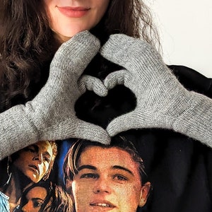 May include: A pair of gray knitted gloves, forming a heart shape. The gloves are made of a soft, textured yarn and have ribbed cuffs. The gloves are a neutral color, suitable for various outfits.