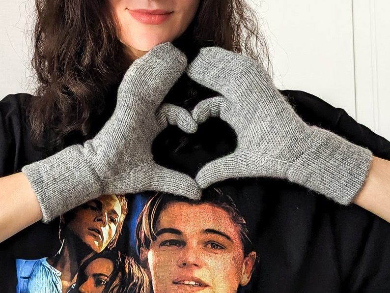 May include: A pair of gray knitted gloves, forming a heart shape. The gloves are made of a soft, textured yarn and have ribbed cuffs. The gloves are a neutral color, suitable for various outfits.