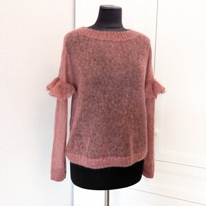 May include: A dusty rose, long-sleeved sweater with a sheer, open-knit texture. The garment features a round neckline with ribbed trim, a matching ribbed hem, and decorative ruffled details at the shoulders where the sleeves attach to the bodice.