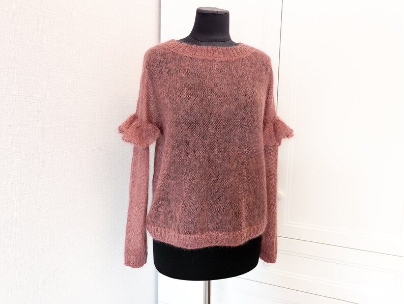 May include: A dusty rose, long-sleeved sweater with a sheer, open-knit texture. The garment features a round neckline with ribbed trim, a matching ribbed hem, and decorative ruffled details at the shoulders where the sleeves attach to the bodice.