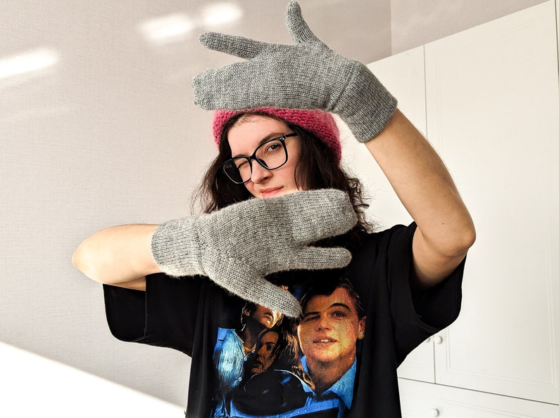 May include: A pair of gray knitted mittens with a matching gray knitted hat. The mittens are made of a soft, textured yarn. The person in the image is wearing a pink beanie, black glasses, and a black t-shirt with a graphic print.