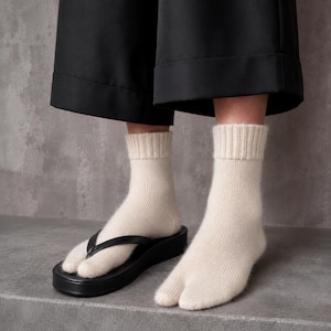 May include: A pair of cream-colored split-toe socks with a ribbed cuff, worn with black thong-style sandals. The socks feature a distinct separation between the big toe and the other toes, designed to accommodate the strap of the footwear.