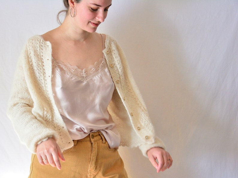 Puede incluir: Rebeca color crema con frente abierto y cierre de botones. La rebeca se combina con una camisola de seda y pantalones color mostaza. La camisola tiene un ribete de encaje. La rebeca est&aacute; hecha de un material suave y texturizado.