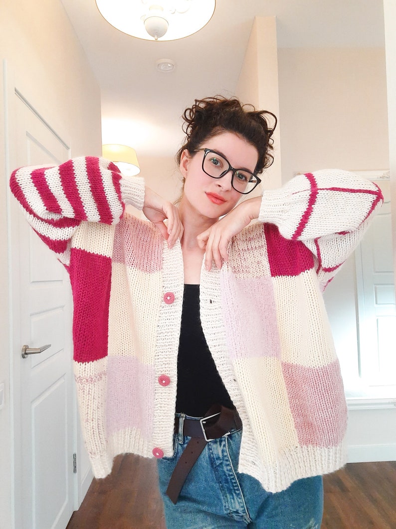 May include: A chunky knit cardigan featuring a color-blocked patchwork design in cream, magenta, and soft pink. The garment has long sleeves, a ribbed collar, and three small pink buttons down the front. It is worn over a black top and blue jeans.