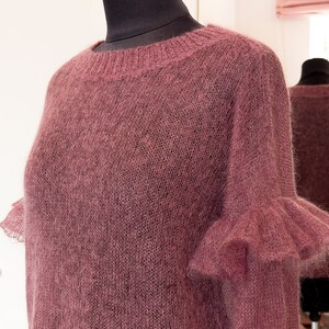 May include: A dusty rose colored long-sleeved sweater featuring a round neckline with ribbed trim and ruffled cuffs at the ends of the sleeves. The garment is crafted from a fine, semi-sheer knit material with a soft, fuzzy texture throughout.