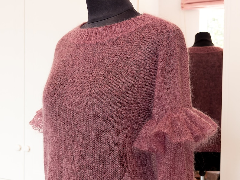May include: A dusty rose colored long-sleeved sweater featuring a round neckline with ribbed trim and ruffled cuffs at the ends of the sleeves. The garment is crafted from a fine, semi-sheer knit material with a soft, fuzzy texture throughout.
