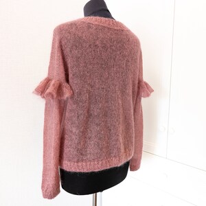 May include: A dusty rose-colored sweater with a sheer, fuzzy knit texture. The garment features long sleeves with ruffled detailing at the upper arms, a round neckline, and ribbed cuffs and hem, displayed on a mannequin torso.