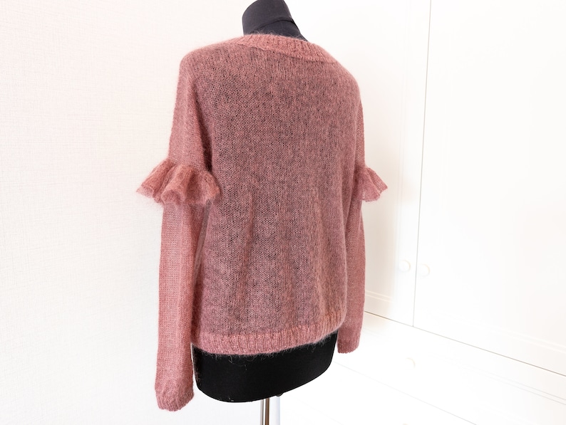 May include: A dusty rose-colored sweater with a sheer, fuzzy knit texture. The garment features long sleeves with ruffled detailing at the upper arms, a round neckline, and ribbed cuffs and hem, displayed on a mannequin torso.