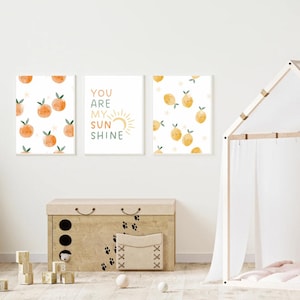 Könnte beinhalten: Drei gerahmte Kunstdrucke mit weißem Hintergrund. Zwei Drucke zeigen Illustrationen von Orangenfrüchten, und der mittlere Druck lautet "YOU ARE MY SUNSHINE" mit einer Sonnengrafik. Eine Holzkiste und ein Spielzelt befinden sich im Vordergrund.