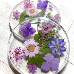 May include: Two round clear resin coasters featuring pressed purple and white flowers. The coasters are ideal for adding a touch of nature to your home decor.