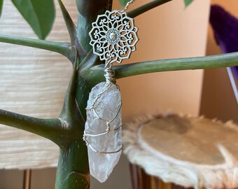Op de afbeelding: Een helder kwartskristal gewikkeld in zilverdraad, hangend aan een zilveren mandala-bedel. Het kristal is bevestigd aan een zilverdraad dat is opgerold in een spiraalvorm.