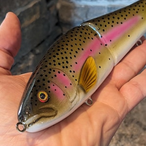 May include: A detailed, realistic fishing lure shaped like a trout. The lure features a yellow, pink, and brown color scheme with black spots. It has a hook and is designed to mimic a real fish.