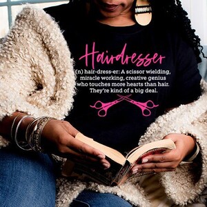 May include: A woman wearing a black t-shirt with the text "Hairdresser (n) hair-dress-er: A scissor wielding, miracle working, creative genius who touches more hearts than hair. They're kind of a big deal." and a pair of pink scissors graphic. She is sitting on a gray couch and reading a book.