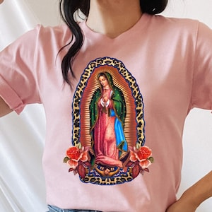 May include: A pink t-shirt with a graphic print of Our Lady of Guadalupe, a religious figure, surrounded by flowers and a leopard print frame.