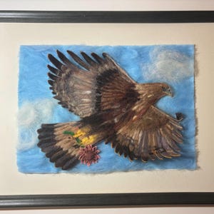 Könnte beinhalten: Gerahmtes Kunstwerk mit einer detaillierten Darstellung eines braunen und tanfarbenen Adlers im Flug vor einem blauen Himmel mit weißen Wolken. Der Adler trägt eine Blume. Das Kunstwerk ist in einem schwarzen Rahmen gerahmt.