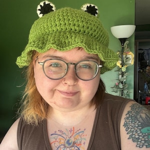 Puede incluir: Un sombrero de cubo de ganchillo verde con ojos de rana. El sombrero tiene un borde con volantes y dos ojos de rana negros y blancos en la parte superior. La persona lleva gafas y una camiseta sin mangas marrón.