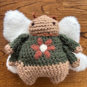 May include: A handmade crochet fairy doll with white fluffy wings. The doll has a brown head, a green body with a flower design, and a tan bottom. The doll is made of yarn and is sitting on a wooden surface.