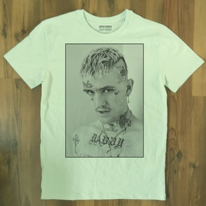 May include: White t-shirt featuring a black and white portrait of a person with tattoos and piercings. The portrait is framed in a black rectangle. The shirt has a crew neck and short sleeves. The text "TEXT SHIRT" is printed on the collar.