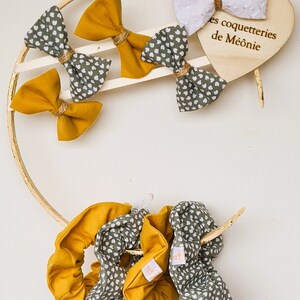 Peut inclure: Un ensemble de chouchous en jaune moutarde et vert avec des motifs de cœurs blancs. Les chouchous sont présentés sur un cercle en bois avec une pancarte en forme de cœur qui indique "Les coquetteries de Méonie".
