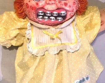 May include: A doll with orange yarn hair tied with a yellow ribbon, wearing light blue glasses and metal braces on its teeth. The doll has a textured, spotted face and wears a yellow dress with white polka dots, a white collar, and matching shorts.