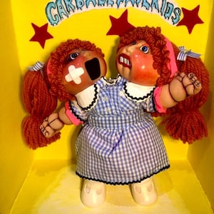 May include: Two Cabbage Patch Kids dolls are joined together at the head, wearing a blue and white gingham dress with a pink collar and black trim. The dolls have red yarn hair and are in a yellow box with the words "Cabbage Patch Kids" written in blue and white.