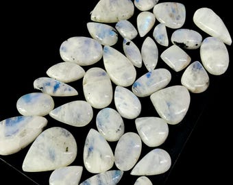 Puede incluir: Una colección de cabujones de piedra lunar pulidos en varias formas, incluyendo ovalados, de lágrima e irregulares. Las piedras son principalmente blancas con sutiles patrones internos azules y grises. El fondo es negro.