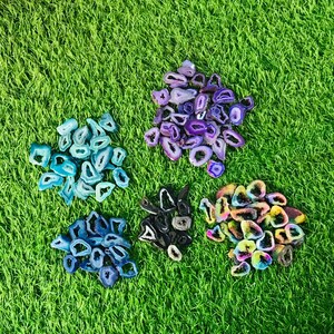 May include: Assortment of agate slices in various colors and patterns, including blue, purple, and rainbow. The slices are arranged on a green grassy surface.