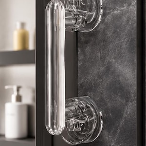 Op de afbeelding: Een cilindervormige deurklink van helder glas met een glad, langwerpig handvat en twee ronde, gefacetteerde montagevoeten. De klink is bevestigd aan een donker, gestructureerd oppervlak en heeft een transparant ontwerp met lichtreflecties.