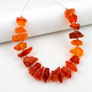May include: A necklace featuring irregular, translucent orange amber-colored stone beads strung on a thin wire. Small, round, metallic silver-toned spacer beads are positioned between each of the larger, uniquely shaped, polished stone segments.