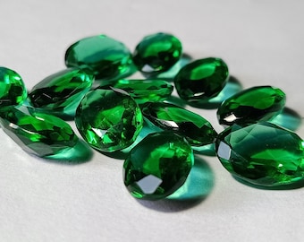 May include: A collection of faceted, emerald-green gemstones. The gems are oval and teardrop-shaped, with multiple facets that catch the light. The stones are scattered on a white surface, creating a sparkling display.