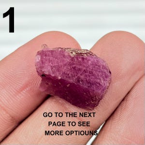 May include: A rough, raw ruby gemstone with a deep pink hue. The stone is held in a hand, with the number "1" in black in the upper left corner. Text at the bottom reads "GO TO THE NEXT PAGE TO SEE MORE OPTIOUNS".