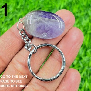 May include: A polished amethyst keychain with a silver chain and key ring. The purple and white stone is oval-shaped. The text "1" is in the upper left corner. The text "GO TO THE NEXT PAGE TO SEE MORE OPTIOUNS" is in the lower left corner.