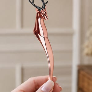 May include: A rose gold-colored metal object shaped like a deer head with black antlers. The deer head transitions into a smooth, curved, tapered handle. The metallic surface is polished and reflective, showing a sleek, elongated, and slender form.