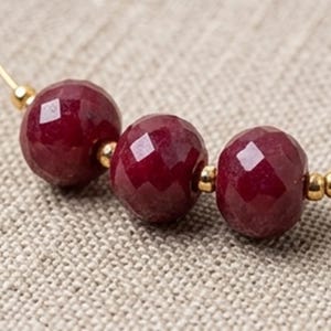 Op de afbeelding: Close-up van een ketting met drie gefacetteerde, dieprode edelsteen kralen. Kleine goudkleurige kralen scheiden de edelstenen, geregen op een dunne gouden draad. De achtergrond is een neutrale, getextureerde stof.