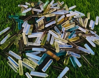 May include: A large pile of various colored crystal points, including clear, white, light blue, and brown. The points are scattered on a bed of green grass.