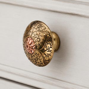 Puede incluir: Un pomo de cajón redondo de latón con un intrincado patrón floral grabado en su superficie. El centro del pomo presenta una incrustación circular texturizada en tono cobre, mientras que el borde exterior tiene un acabado en latón antiguo pulido.