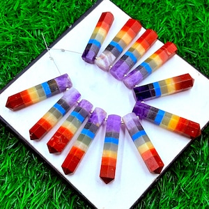 May include: A collection of colorful, faceted gemstone pendants arranged on a white surface. Each pendant features a spectrum of colors, including red, orange, yellow, green, blue, and purple, representing the chakras. The pendants are strung on a silver wire.
